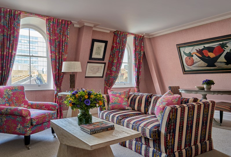 A lounge area with a fora and armchair facing a coffee table in the center, dining table for 2 at the rear and 2 windows streaming light in to the side. Colourful artwork hangs above the table and between the windows.
