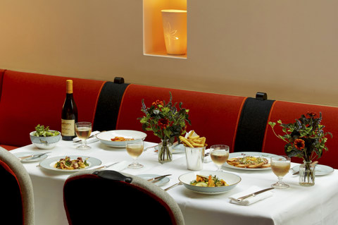An image to promote the à la carte mains at Ham Yard Bar & Restaurant at Ham Yard Hotel. The image shows a white-linen table set with a selection of starters, including oysters, prawn cocktail, and vibrant salads. There are wine placed next to each dish, set against a backdrop of Martha Freuds ceramic installation. 