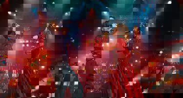 A still from 'Song Sung Blue'. A man dressed in a sparkling blue outfit and a woman dressed in a sparkling red outfit stand opposite one another singing on stage with a large band behind them