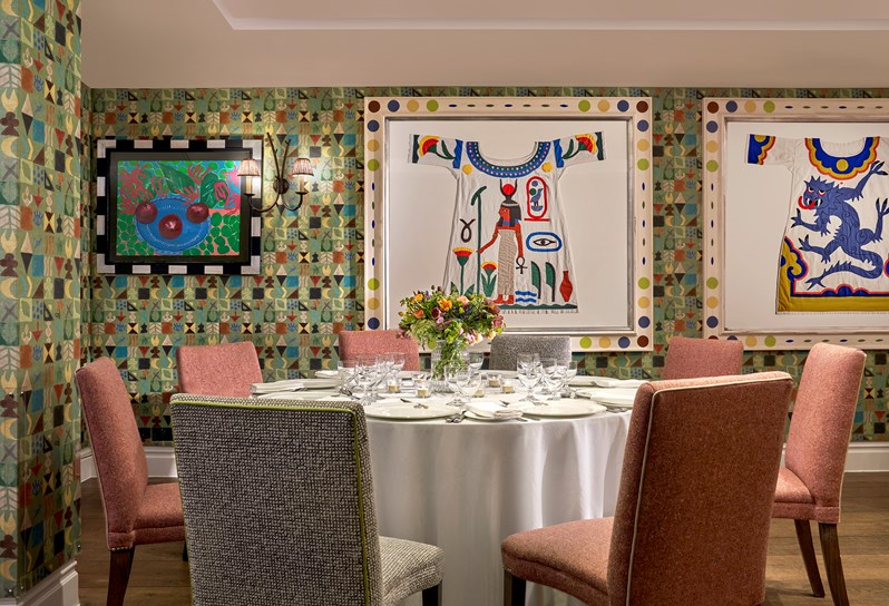 A closeup of a table in the newly photography Vanessa Bell Room setup for a dinner orange chairs. There is beautiful bright artwork on the walls and flowers decorating the table.
