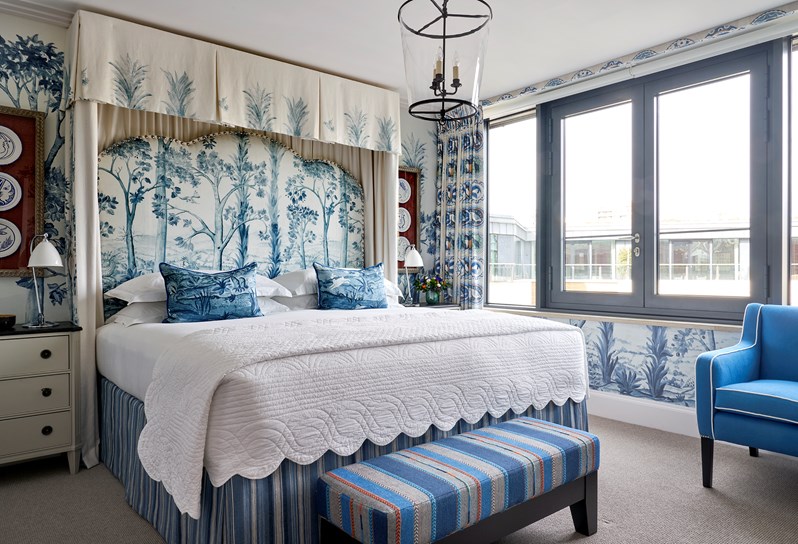 A canopied bed with a blue patterned headboard with blue cushions and a white valance. A blue and orange patterned footstool sits at the end of the bed