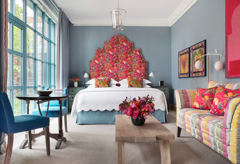 A blue-walled bedroom featuring a double bed with a pink patterned headboard and cushions and two love heart prints on the wall to the right. In front there is a small table with two blue chairs and a multi-coloured striped sofa opposite a wooden coffee table.