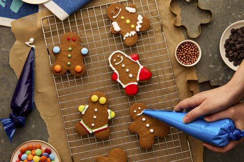 This image shows festive gingerbread decorating to promote festive dining at Brumus, Haymarket Hotel, with colourful gingerbread people arranged on a cooling rack alongside icing bags, sweets and sprinkles, as a gingerbread biscuit is decorated by a blue icing bag.