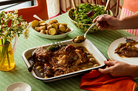 An oven dish filled with slow cooked lamb placed on a table with a hand taking a spoonful out to put on a nearby plate. The lamb is accompanied by potatoes and salad