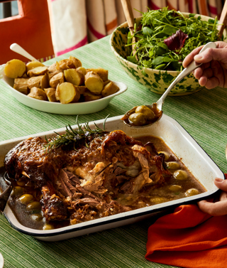 An oven dish filled with slow cooked lamb placed on a table with a hand taking a spoonful out to put on a nearby plate. The lamb is accompanied by potatoes and salad