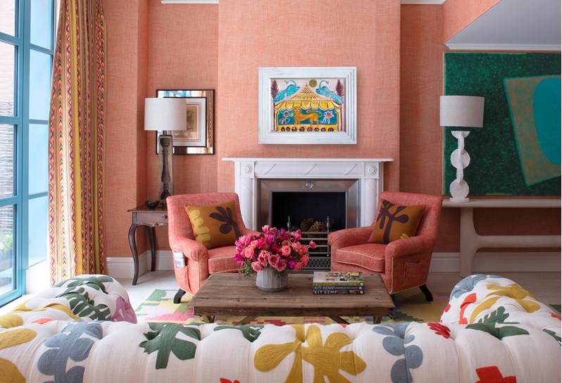 A living room space with orange walls, a multicoloured sofa and orange chairs. At the end of the room there is a fireplace above which hangs a piece of art featuring a folk art inspired lion