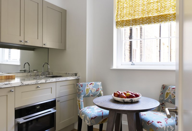 A kitchen with marble countertop, big window with yellow curtain and two chairs in blue patterned fabric with table.