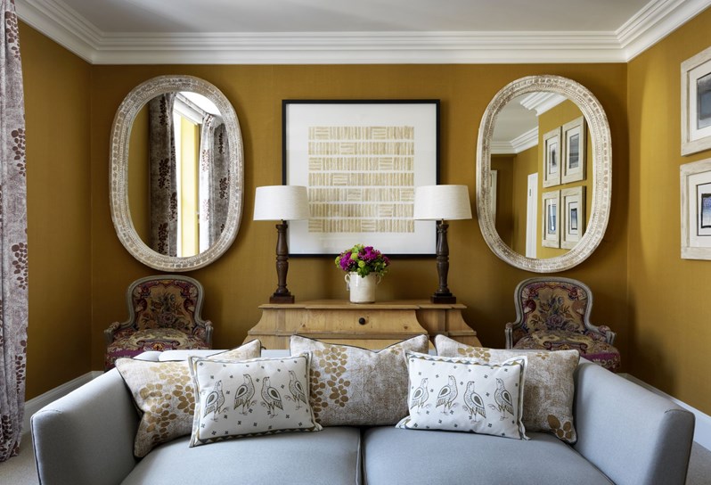 The living space of a Suffolk suite in warm gold tones with a neutral couch with decorative pillows. Behind are two mirrors framing a piece of art and two chairs.