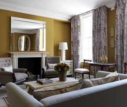 The living room to a Suffolk suite in a warm gold yellow tone. The sitting area has a couch and two chairs in more neutral tones in front of a fireplace with a large mirror and patterned curtains on two large windows.