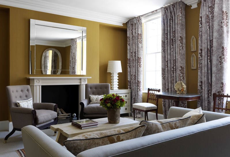 The living room to a Suffolk suite in a warm gold yellow tone. The sitting area has a couch and two chairs in more neutral tones in front of a fireplace with a large mirror and patterned curtains on two large windows.