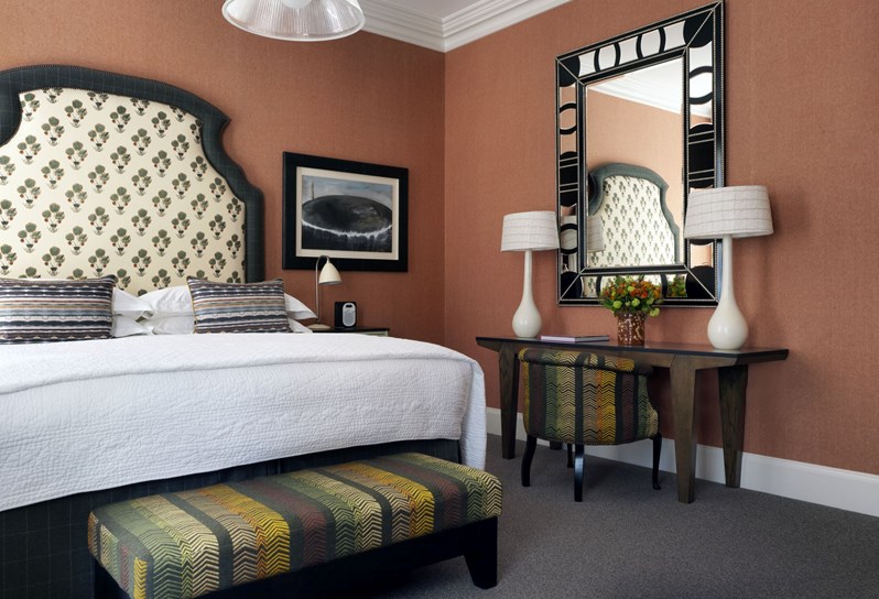 A bedroom featuring burnt orange walls, a cream patterned headboard with decorative throw pillows and a multicolored end of bed footstool. To the right there is a writing desk with a large mirror and flowers.