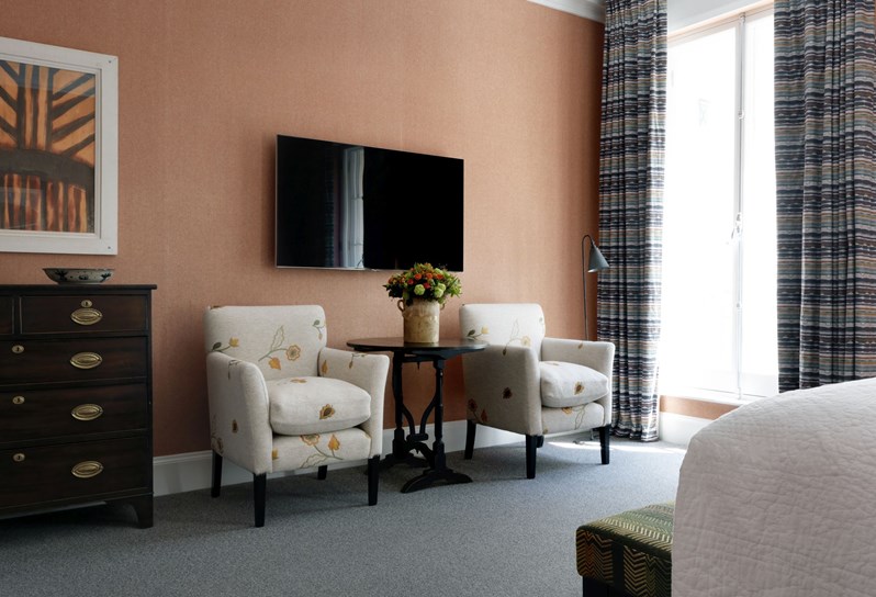 The seating area of a bedroom with burnt orange walls, chairs in a cream fabric with flowers on them, a television and horizontally striped curtains.