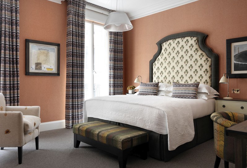 A bedroom featuring burnt orange walls, a cream patterned headboard with decorative throw pillows and a multicolored end of bed footstool. There is a cream floral patterned chair in front of the windows which have horizontally striped drapes.