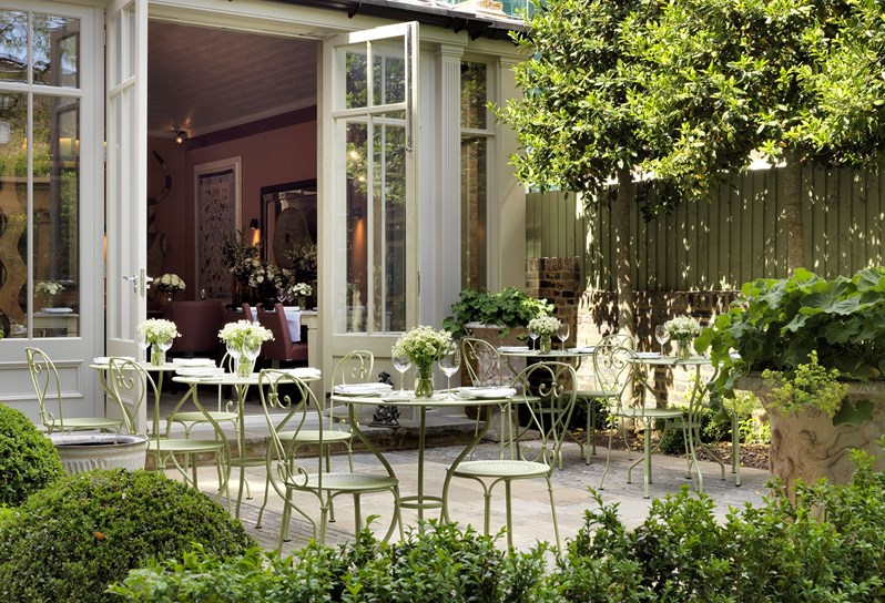 The Garden at Number Sixteen looking back towards the hotel's Orangery where the doors are open.  Green planting and a patio showing 5 sets of wrought iron chairs and tables decorated with white and green flowers and set for dining.