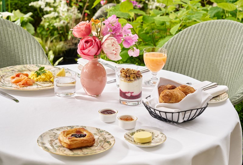Table outside with two wicker chairs surrounded by plants. On the table there is a bowl of pastries, a pot of yoghurt and granola, a plate with scrambled eggs and smoked salmon, a plate with a jam pastry on and side dishes with butter and jams. There is also a glass of water and juice and bud vase of pink flowers.