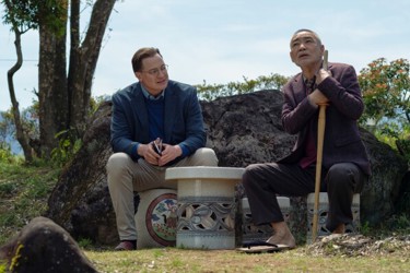 Still from 'Rental Family'. A smartly dressed white man sits on a stool next to an older Asian man grasping a wooden staff. They are surrounded by rocks and greenery