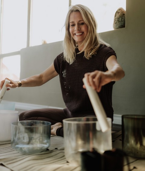 A close up shot of sound practitioner Nancy Trueman sitting on the floor playing crystal bowls with two large sticks