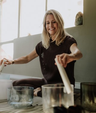 A close up shot of sound practitioner Nancy Trueman sitting on the floor playing crystal bowls with two large sticks