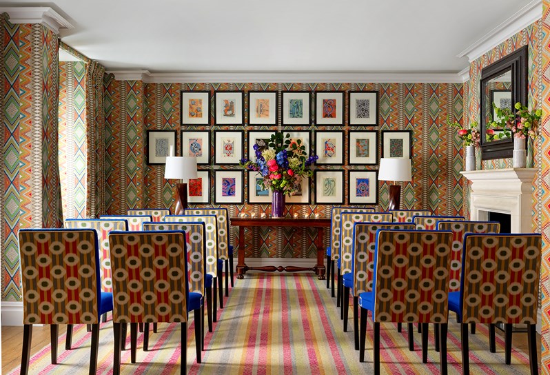 A room is set up for a wedding ceremony with chairs placed either side of an aisle and a large vase of flowers on a table at the end