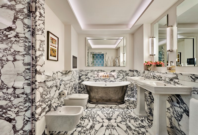 A black and white marble tiled bathroom with a large metal bathtub and a white toilet, bidet and two twin sinks, plus 3 mirrors on the wall