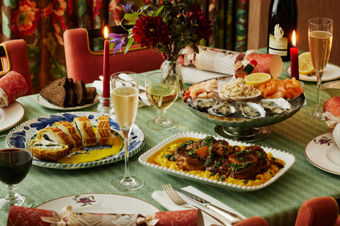 This image show the Christmas Eve dining menu at Refuel, The Soho Hotel, with a festive table set with Mythical Creatures crockery. It features a seafood platter of oysters, prawns, smoked salmon and dressed crab, Sea bass en croûte, spinach and Champagne beurre blanc, alongside Osso buco, saffron risotto and gremolata. Champagne, wine, candlelight, seasonal flowers and Christmas crackers complete the setting.
