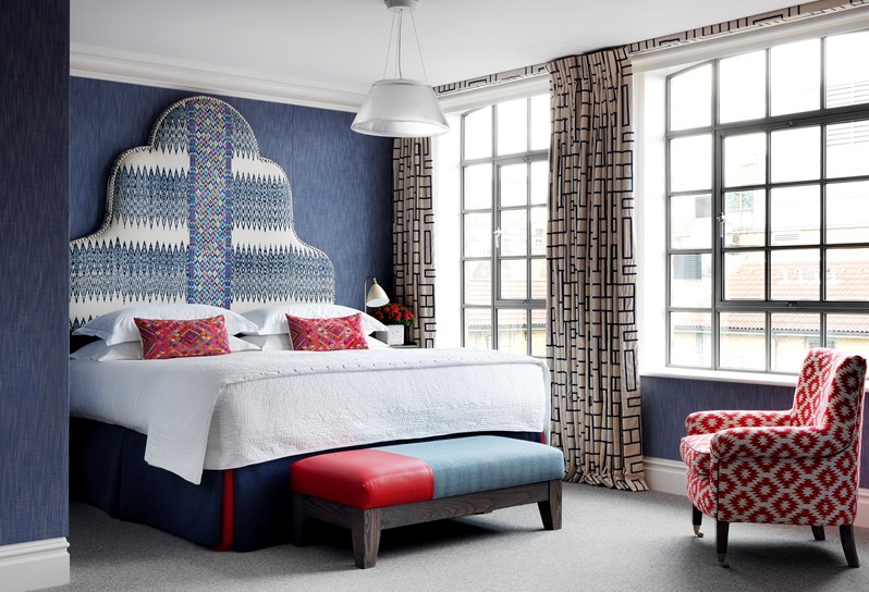 A bedroom with multi coloured headboard, dark blue valance and pink and red pillow on the bed. At the foot of the bed is a red and blue ottoman. A patterned red and white armchairs faces the bed. Black and white curtains frame the windows, which stream light in.