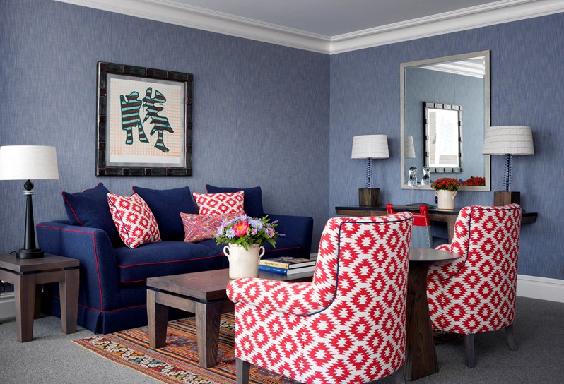 An image of the sitting area in one of the Deluxe Junior Suites. There is a large blue sofa with bold red piping and white and red decorative pillows. The walls are a muted, soft blue and a large modern painting with a metal frame hangs on the wall. Two red and white arm chairs face the coffee table, which sits on a multicolored rug. A large mirror hanging on the wall and a side table faces the sitting area with two designer lamps.