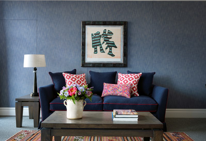 An image of the sitting area in one of the Deluxe Junior Suites. There is a large blue sofa with bold red piping and white and red decorative pillows. The walls are a muted, soft blue and a large modern painting with a metal frame hangs on the wall. The large wooden coffee table in the center sits on a multicolored rug. A side table is on the right of the sofa with a designer lamp with a black base and white lampshade.