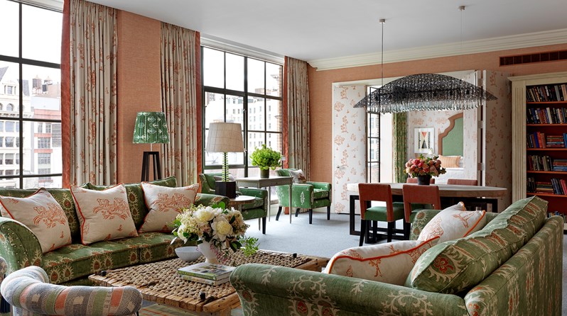 A spacious Crosby Suite with green and orange colour scheme. There are two green sofas surrounding a coffee table. In the background is a dining table. The suite has floor length windows over looking the SoHo neighbourhood.