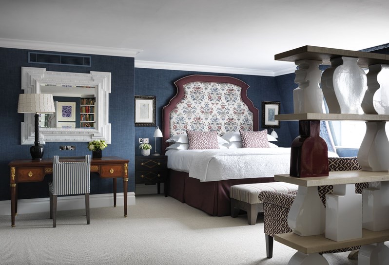 The Penthouse Suite bedroom with blue walls and a burgundy outlined headboard on the bed. There is a desk to the left with a large mirror hanging above and shelving to the right of the room.