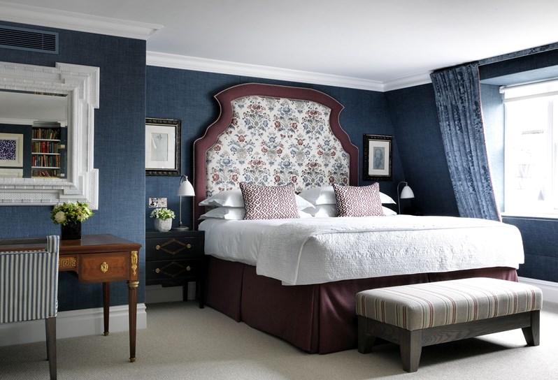 The Penthouse Suite bedroom with blue walls and a burgundy outlined headboard on the bed. There is a desk to the left with a large mirror hanging above and a window to the right.
