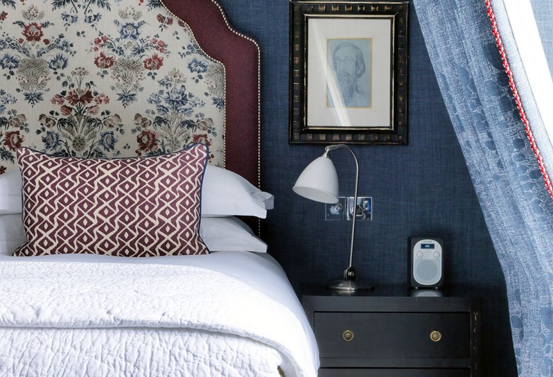 The Penthouse Suite bed with a floral burgundy outlines and floral headboard takes up this image on the left with blue curtains on the right.