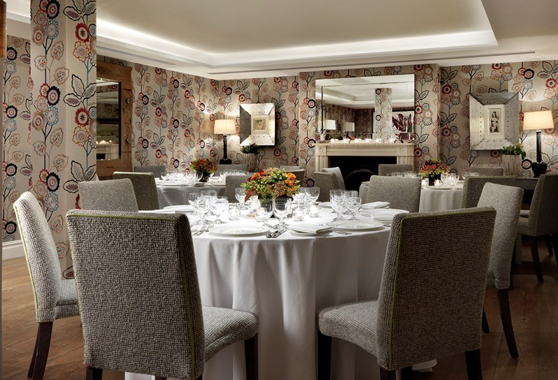 The Duncan Grant Room setup for a dinner on round tables. The walls are decorated with a beautiful floral wallpaper and there is a large mirror hanging above a fireplace at the back of the room.