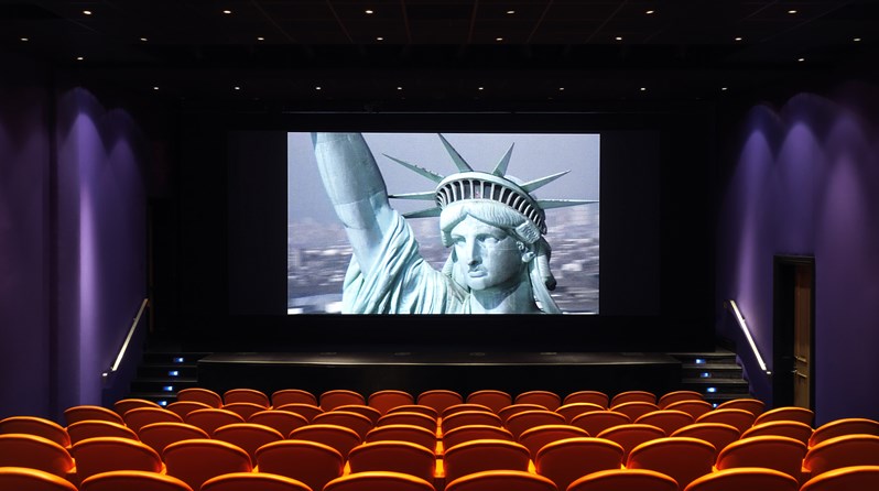 The screening room at the Crosby Street Hotel has 99 red leather seats by the Italia furniture company Poltrona Frau. there is a stage and wide screen here pictured with an image of the Statue of Liberty.