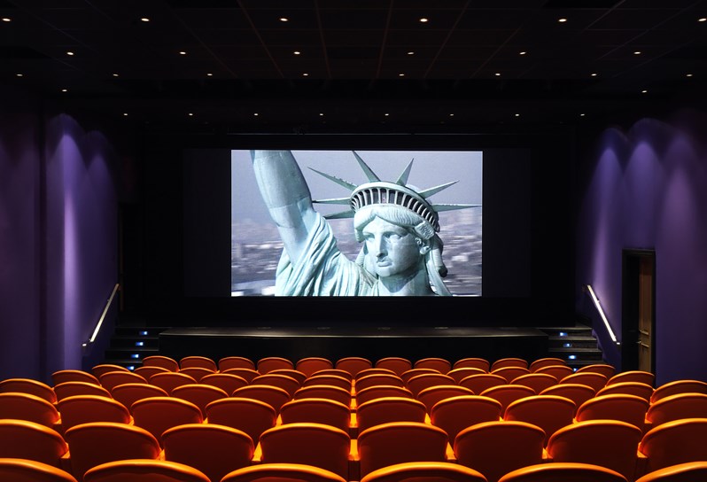 The screening room at the Crosby Street Hotel has 99 red leather seats by the Italia furniture company Poltrona Frau. there is a stage and wide screen here pictured with an image of the Statue of Liberty.
