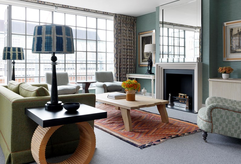A colorful living room with green couch facing the fireplace. An orange rug sits under the coffee table. Lamps with patterned blue and white shades sit on either side of the couch. To the back are the floor to ceiling windows and a door leading out onto the terrace.