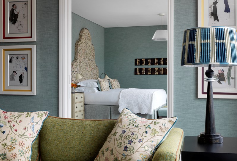 A close up view looking from the living room into the bedroom. Light blue walls are accented by colorfully embroidered pillows and headboard. In the forefront is the corner of a green couch and a lamp with blue and white patterned lampshade. Art work frames both sides of the entry way to the bedroom.