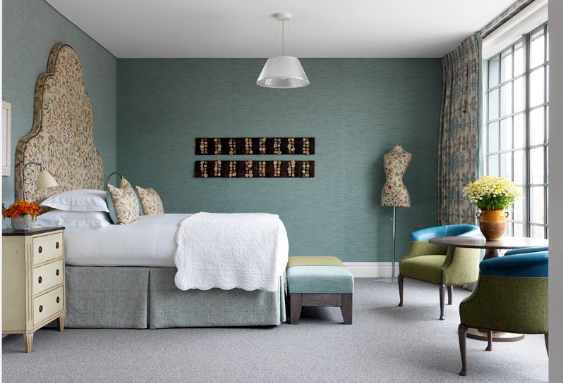 Side view of a bedroom with light blue walls. Neutral fabric with colorful stitching covers the headboard, pillows and mannequin. In front of the windows is a sitting area with two chairs in green and blue and a table with bright yellow flower arrangement.
