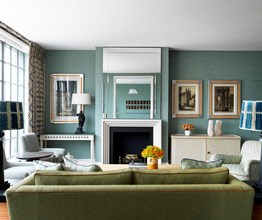 A view from behind a green couch of a living room facing the fireplace.  Lamps with patterned blue and white shades sit on either side of the couch. Above the fireplace is a mirror in which two horizontal art pieces are reflected. There are two flower arrangements in orange, one on the coffee table in front of the couch and one on the table next to the fireplace.