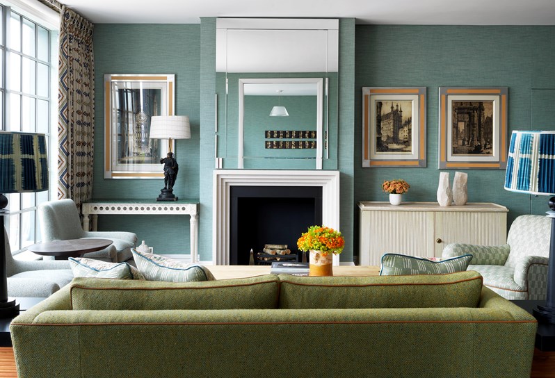 A view from behind a green couch of a living room facing the fireplace.  Lamps with patterned blue and white shades sit on either side of the couch. Above the fireplace is a mirror in which two horizontal art pieces are reflected. There are two flower arrangements in orange, one on the coffee table in front of the couch and one on the table next to the fireplace.