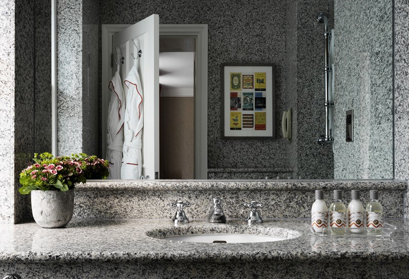 A basin in a granite bathroom, with a plant on the left and four products from Kit Kemp's own bathroom range, RikRak, to the right, as well as a large mirror on the wall showing two fluffy bathrobes hung on the back of a door.
