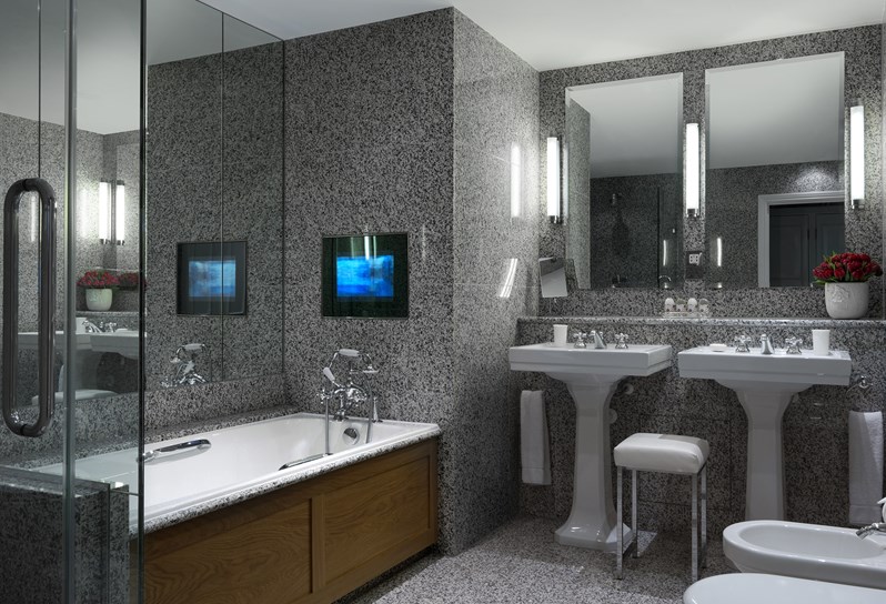 A beautiful granite bathroom with walk in shower, bathtub and television to the left with two wash basins in the center