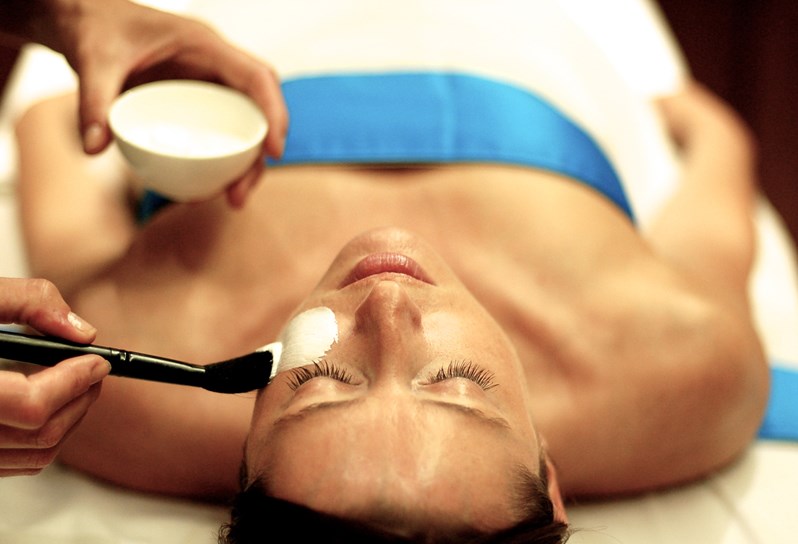 Woman lying on her back on a massage bed having a facial done with a white face mask by a therapist.