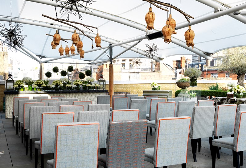 The roof terrace at Ham Yard Hotel set for a wedding ceremony with rows of seats facing forward. Wicker lanterns hang overhead.