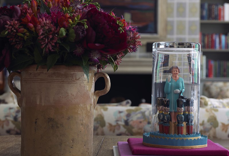 Ceramic vase with fresh flowers in deep reds and purples placed next to a quirky piece of art showing the Queen and some of her Beefeater's encapsulated in a glass goblet on a table in the Library.