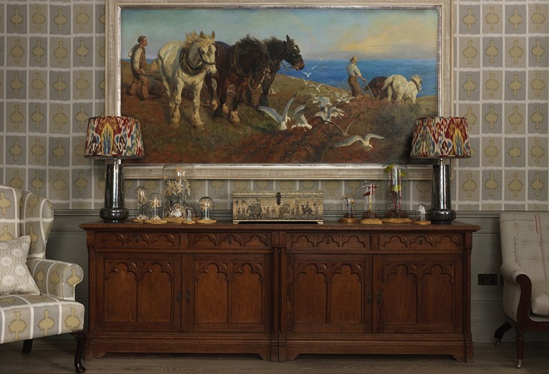 A large painting of horses in nature hung above a large wooden cabinet with quirky artwork and designed lamps placed on top and two comfortable armchairs on either side of the cabinet.