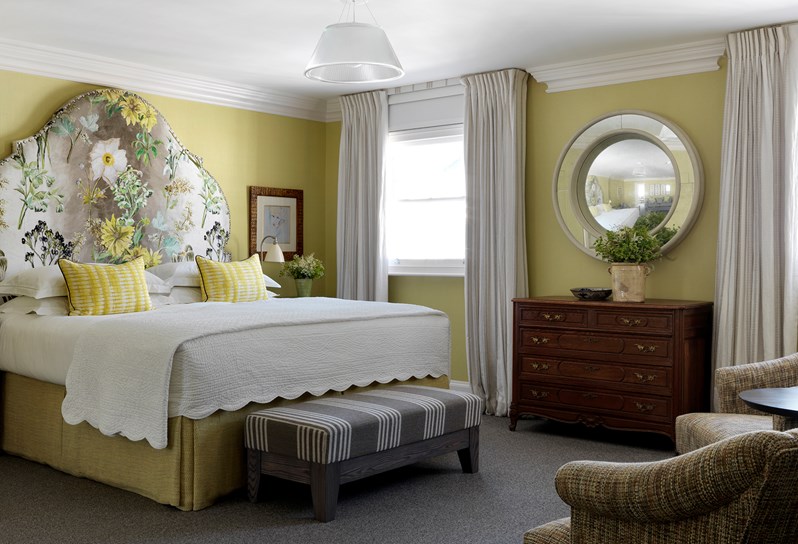 A bedroom in light shades of yellow, white and green with a beautiful floral printed headboard. There are yellow throw pillows and an end of bed footstool in a white and brown striped fabric. Flowers sit on a dresser with a circular mirror on the wall behind it.