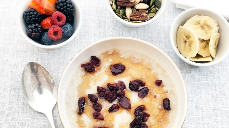 Over head image of a bowl of porridge with honey and dried cranberry porridge. On the table are also small bowls of berries, nuts and sliced bananas.