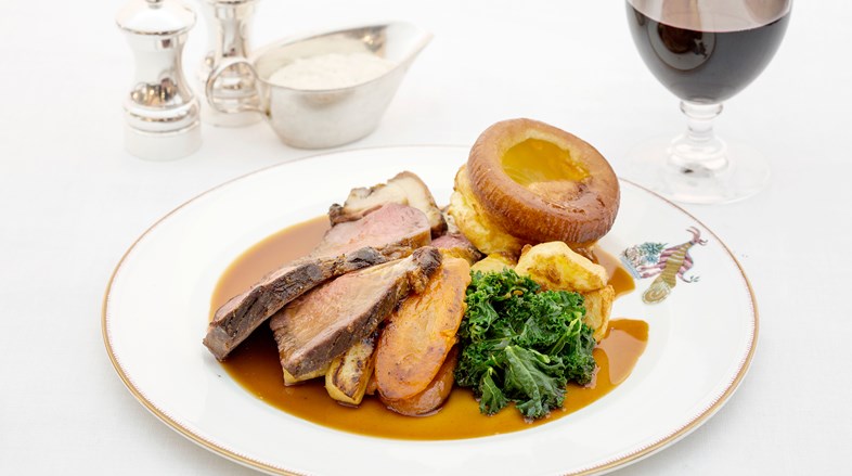 Close up image of a Sunday roast with roast beef, potatoes, gravy and kale. There is a glass of red wine beside the plate.
