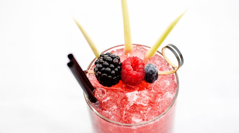 A bright pink cocktail with crushed ice and garnished with a blackberry, raspberry and blueberry on a cocktail stick.